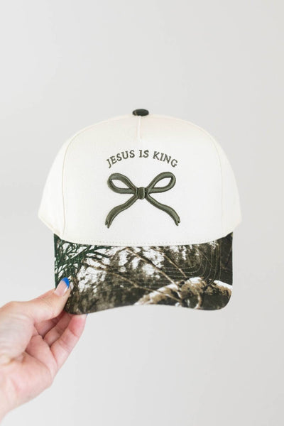 White cap with 'Jesus is King' text and bow design, held by a hand against a light gray background.