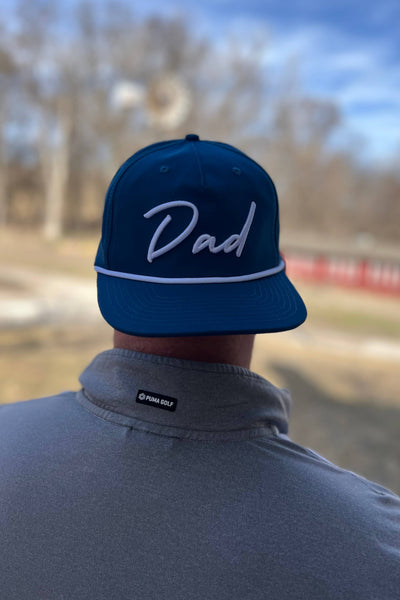 Person wearing a blue cap with 'Dad' on it, standing outdoors with trees and sky in the background.
