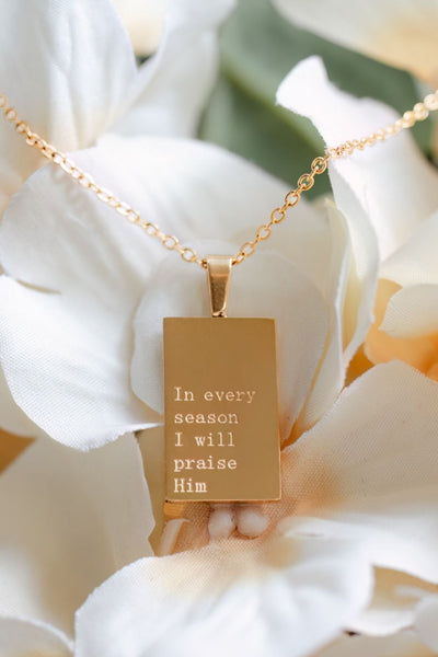 A gold rectangular pendant necklace with engraved text reading 'In every season I will praise Him,' displayed on a delicate gold chain over large white flowers.