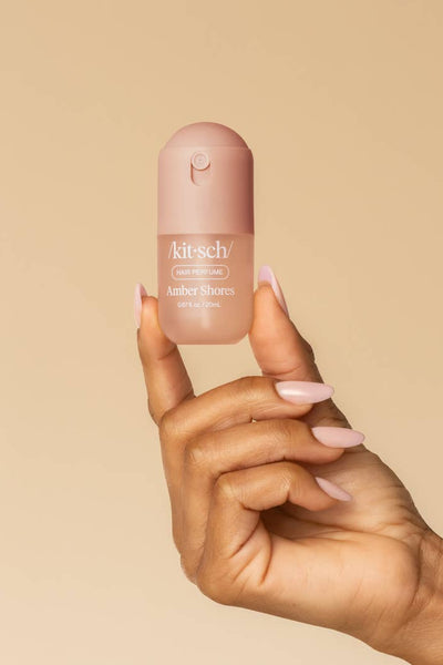 A hand with pale pink manicured nails holds a small bottle of Kitsch Cosmetics liquid shimmer against a solid beige background.