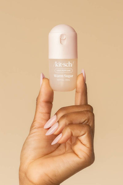 A hand with pale pink manicured nails holds a small Kit-schi Warm Sugar bottle against a solid warm beige background.