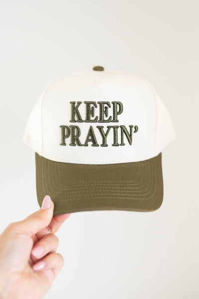 Trucker hat with 'KEEP PRAYIN'' text held by a hand against a white background