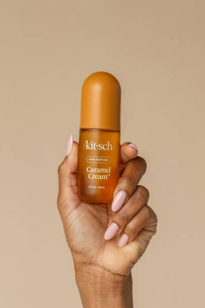 A hand with light pink manicured nails holds a small amber bottle of kitsch Caramel Gloss in front of a solid beige background.