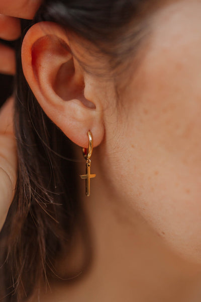 Close-up of an ear wearing a gold hoop earring with a cross pendant.