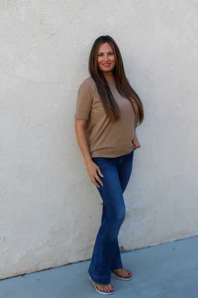 Woman wearing a brown shirt and blue jeans standing against a white wall.