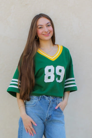 Woman wearing a green jersey with the number 89 and blue jeans against a beige wall.