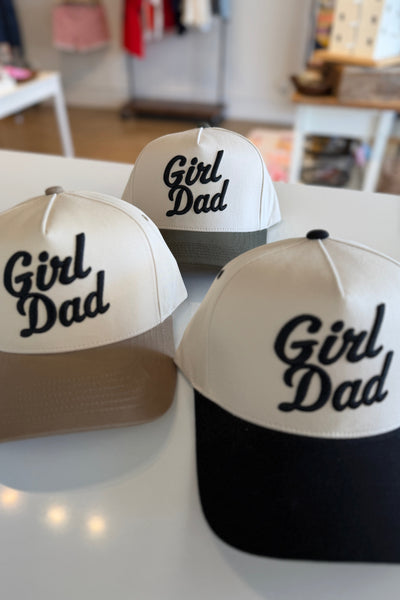 Three 'Girl Dad' hats displayed on a table with a blurred indoor background.