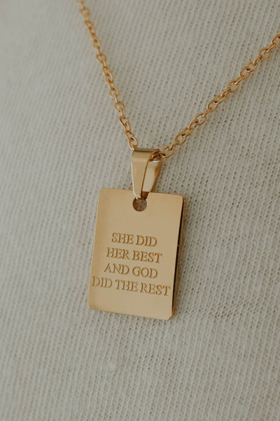 A gold rectangular pendant necklace engraved with the words 'SHE DID HER BEST AND GOD DID THE REST', displayed on a textured off-white fabric background.