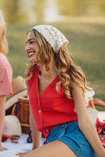 Two women sitting outdoors with a picnic blanket and drinks, one wearing a red shirt and denim skirt.