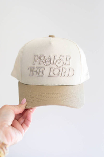 Beige cap with 'PRAISE THE LORD' text held by a hand on a white background