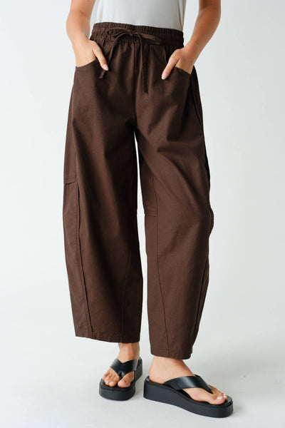 A person wearing loose brown drawstring pants with side cargo pockets, paired with black platform sandals, shown from the waist down against a light gray studio background.