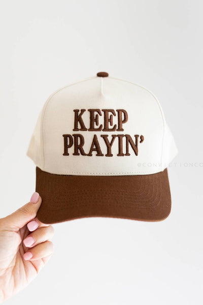 Cap with 'KEEP PRAYIN'' text held by a hand on a white background