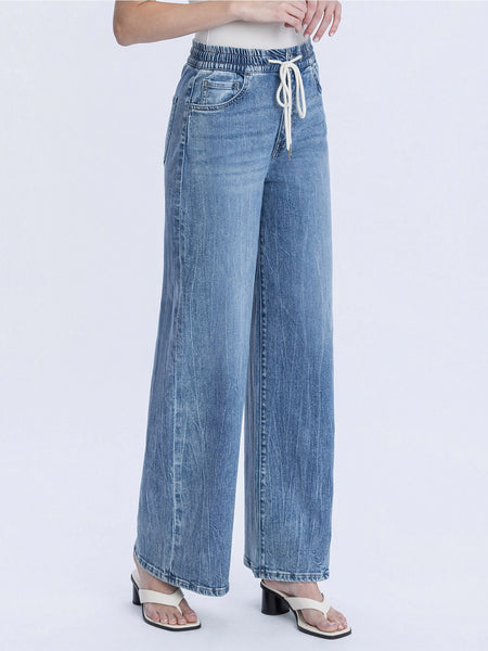 A person wearing light blue wide-leg denim jeans with an elastic waistband and white drawstring, paired with white heeled sandals against a plain light gray studio background.