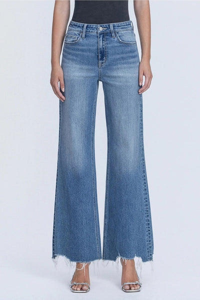 A model wearing a pair of wide-leg, high-waisted blue denim jeans with a raw frayed hem, paired with silver heeled sandals against a light gray studio background.
